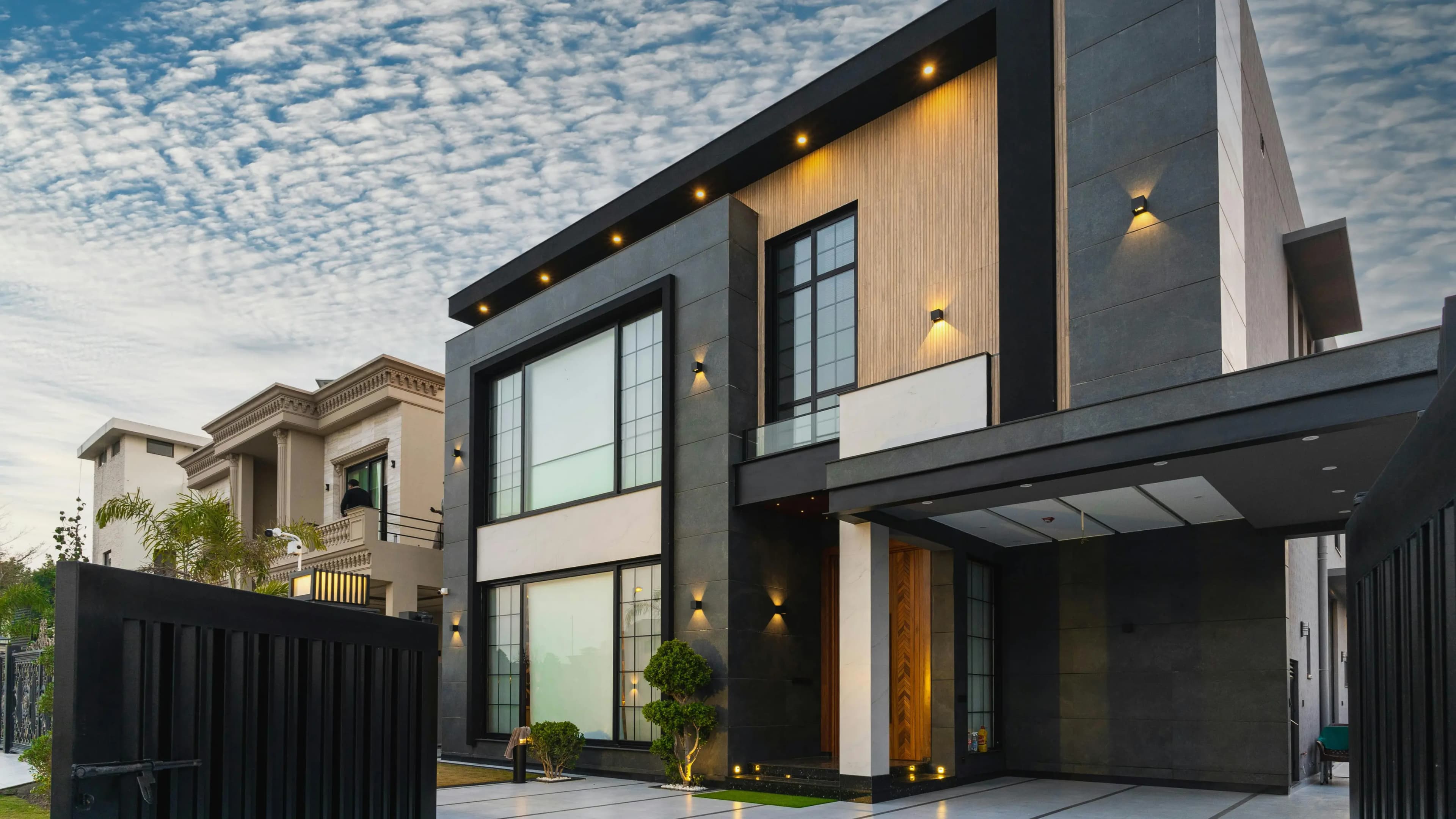 Modern two-storey house with a dark stone facade, warm wall lights and a covered driveway at dusk